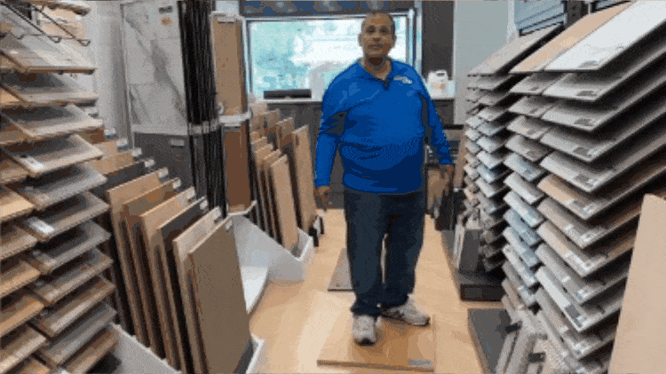 A person stands in a flooring store, surrounded by vertical racks displaying various floor samples. The individual is wearing a blue shirt and jeans, and the store appears well-lit with a window in the background.
