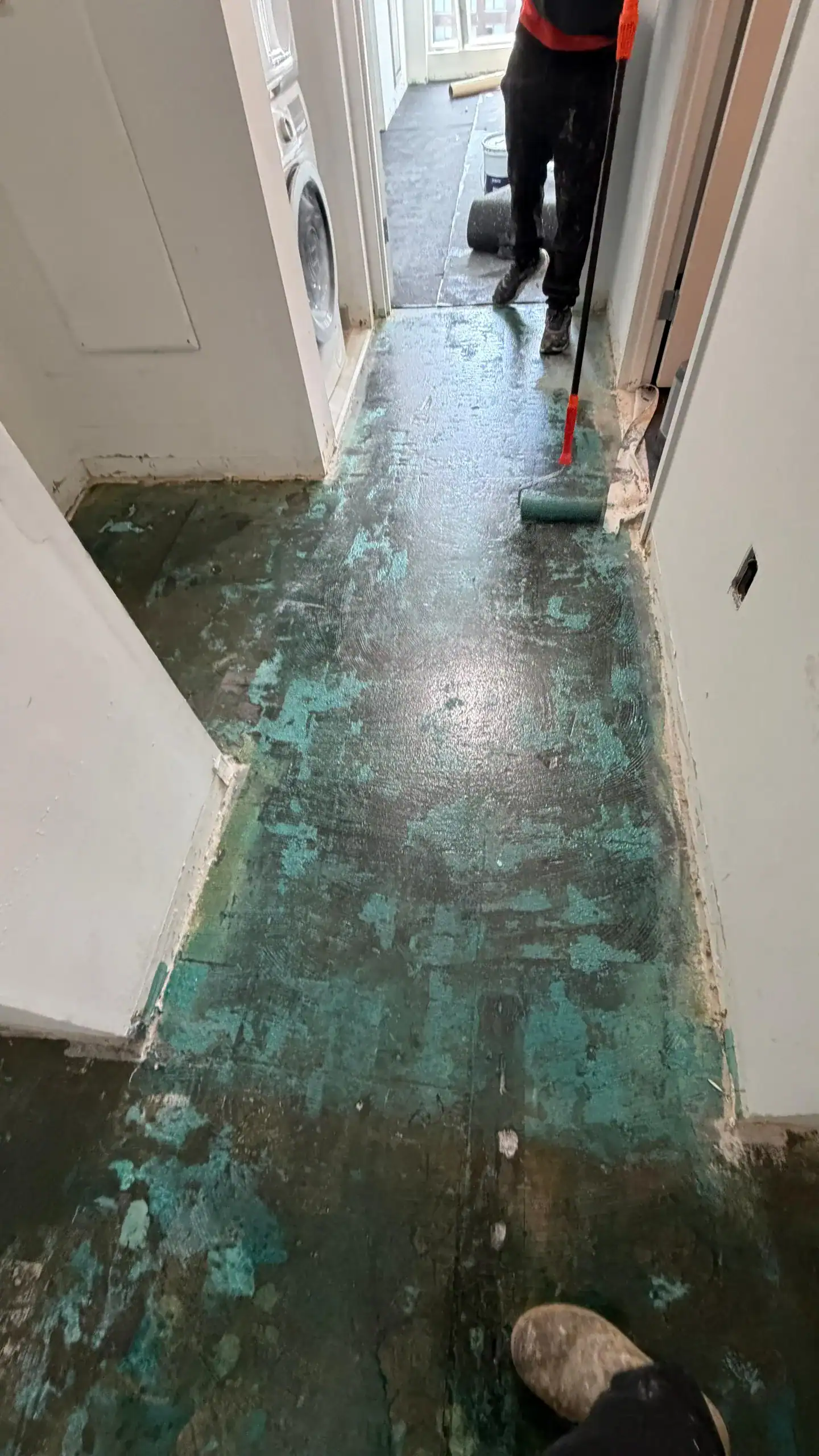 A person uses a roller to apply a coating or adhesive to a hallway floor with worn, patchy, greenish-blue and black areas. Construction or renovation work is in progress. The hallway leads to other rooms.