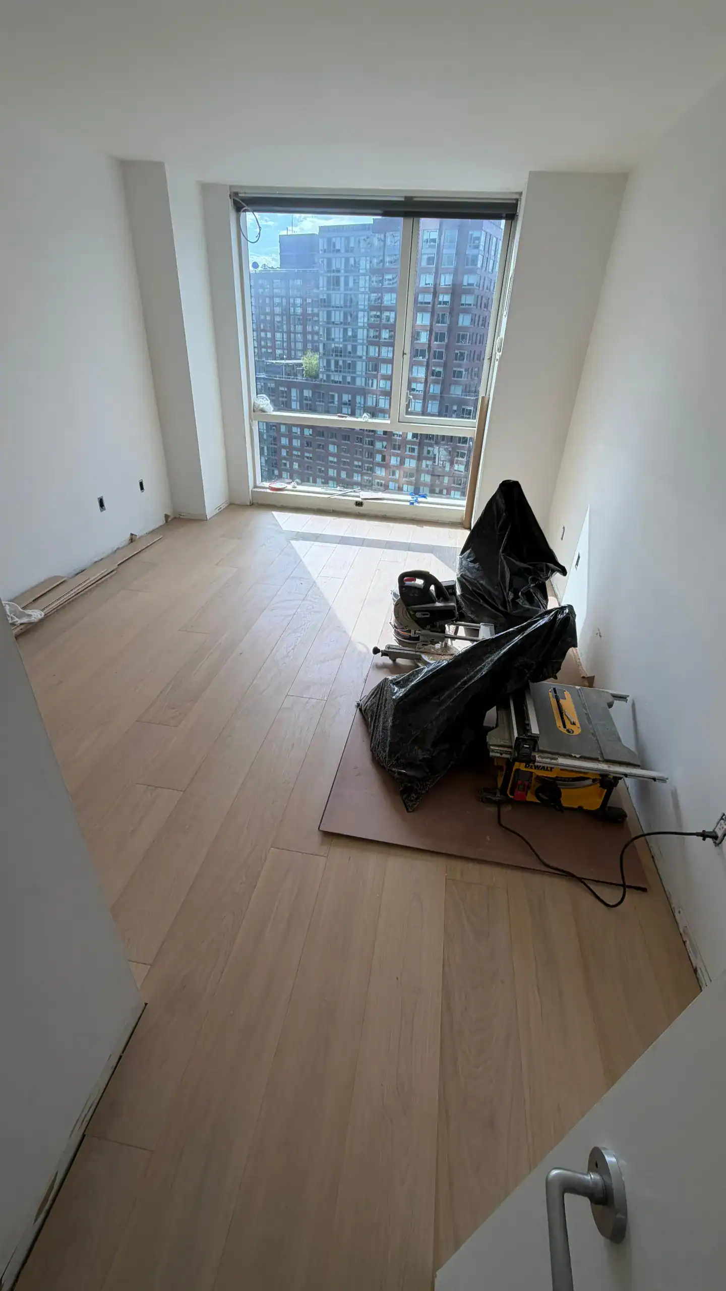 A small, unfurnished room with light wood flooring under construction. Power tools and materials are on the floor near a large window, which offers a view of city buildings outside.