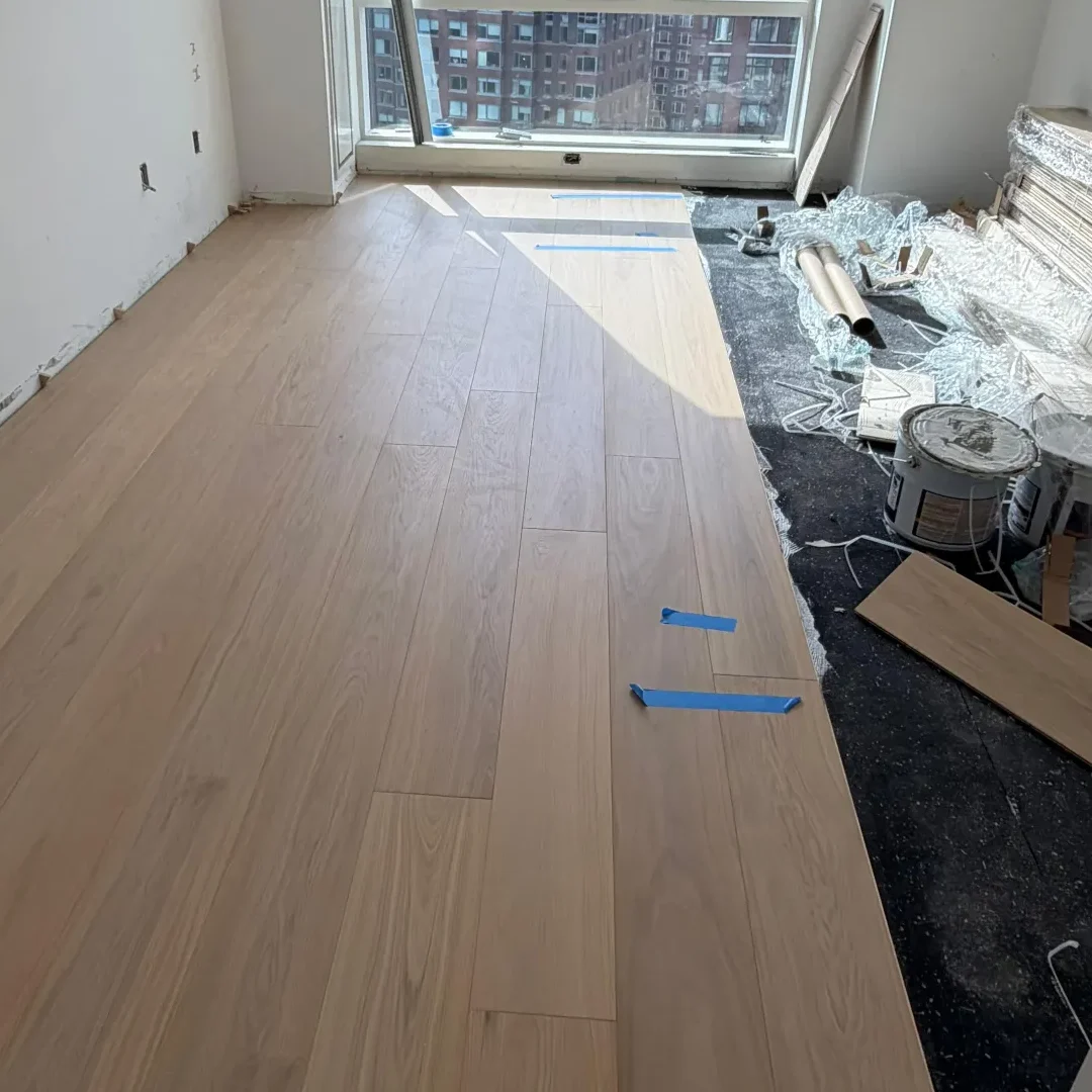 A room under renovation with new light engineered wood flooring partially installed, tools and construction materials on the right, and large windows showing a cityscape of tall apartment buildings outside.