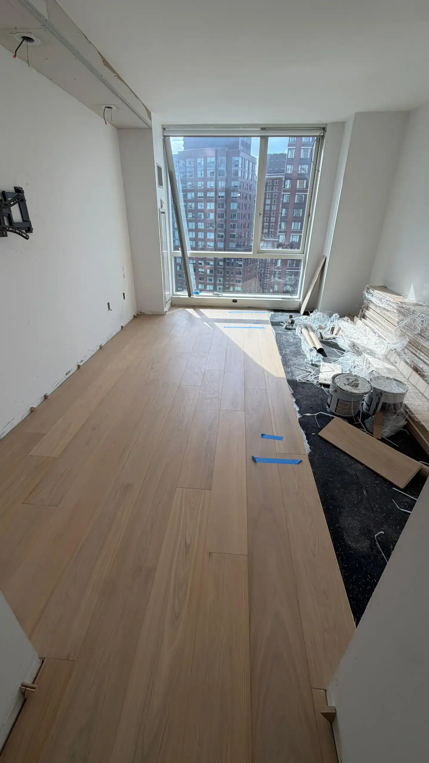A room under renovation with new light wood flooring partially installed, tools and construction materials on the right, and large windows showing a cityscape of tall apartment buildings outside.