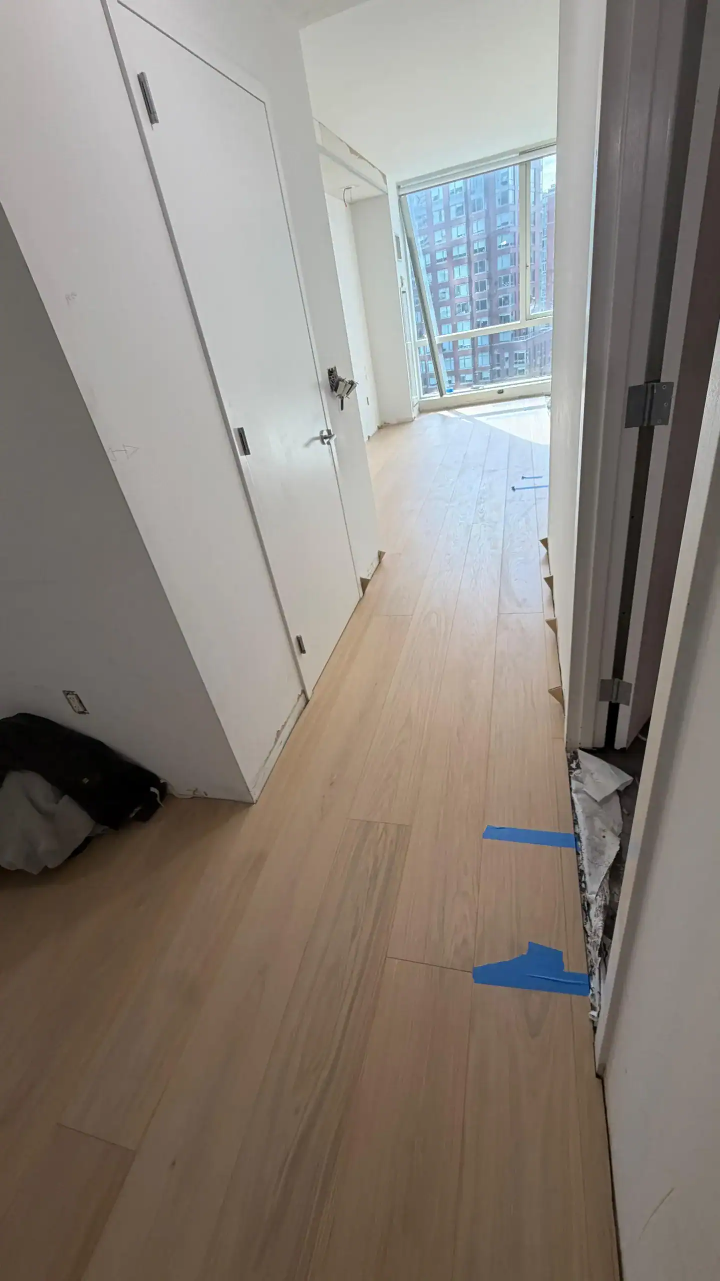A hallway with light wood flooring leads to a bright room with large windows. Blue tape marks a spot on the floor near an unfinished area with exposed material behind an open door. A jacket lies on the floor by a closet.