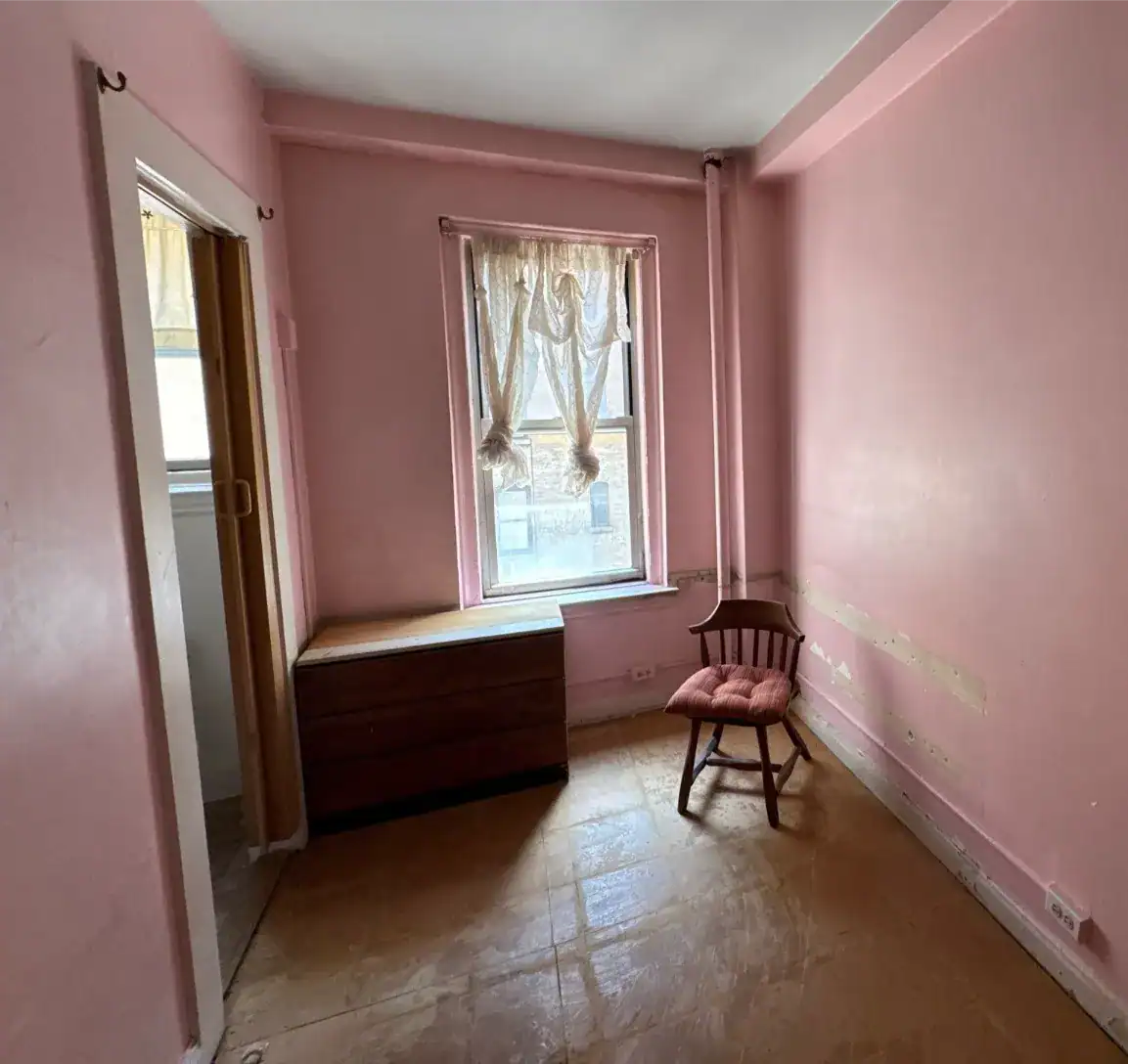 A small, empty room with pink walls, a single window with sheer curtains, a wooden chest under the window, and a wooden chair with a pink cushion. Light streams in, illuminating the bare, worn floor.