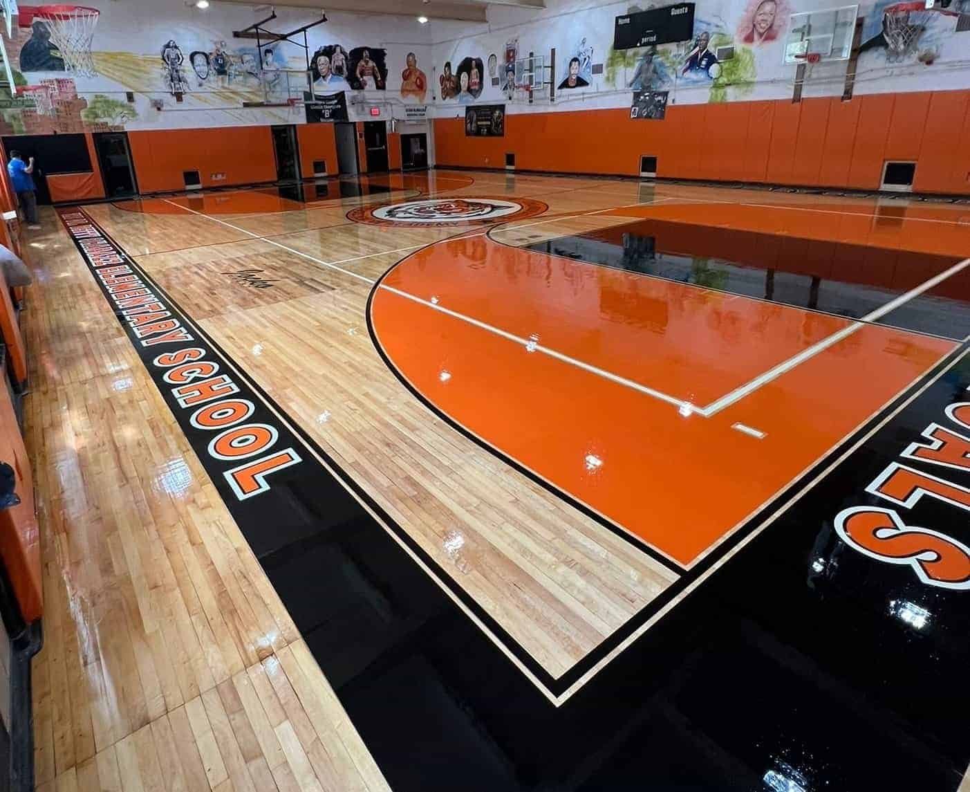 Newly polished school gymnasium with orange and black basketball court, wall murals, hoops, and bleachers. Elementary School text is visible on the floor near the edge of the court.